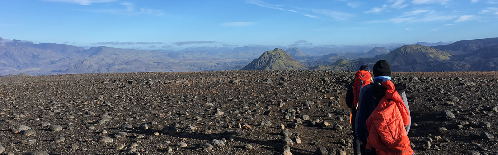 Two individuals hiking across a flat, desolate landscape of dark rocks and gravel. Jagged mountains are close on the horizon.